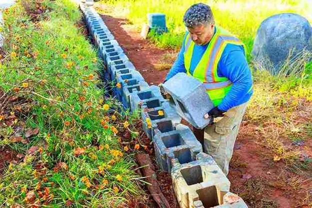 Worker stacking large concrete blocks to build a sturdy retaining wall along a landscape.