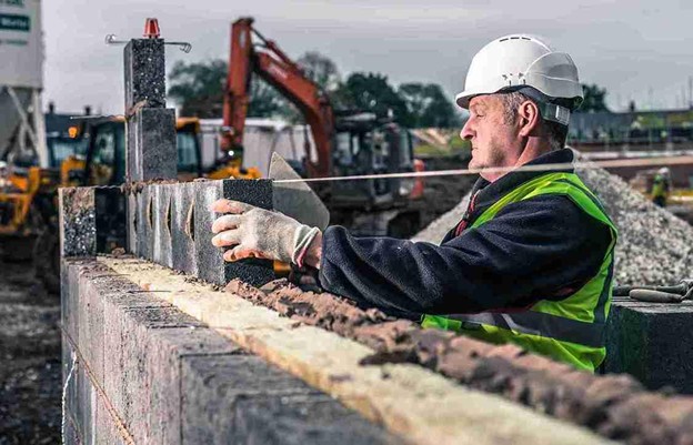 Builder placing concrete blocks to construct a retaining wall on a job site with heavy machinery in the background.